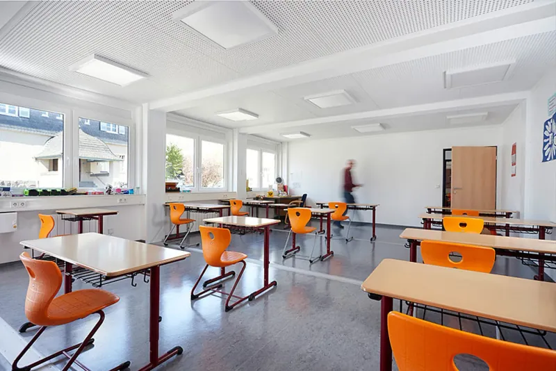 Heller Klassenraum der Grundschule Eibach mit großen Fenstern, Holztischen und orangefarbenen Stühlen